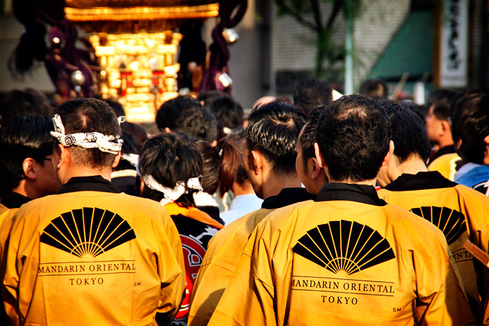 Una processó al centre de Tokyo, la gent estan d'esquena i van tots vestits de color daurat, i porten escrit a l'esquena: Mandarin Oriental Tokyo