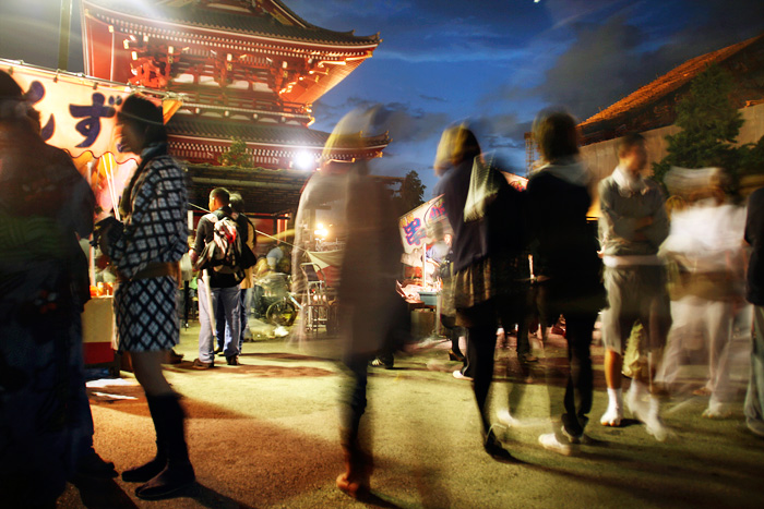 El temple d'Asakusa, al capvespre, un dia de celebració. Hi ha gent vestida a la manera tradicional, uns quiets, altres es mouen, i hi ha algunes paradetes de menjar.
