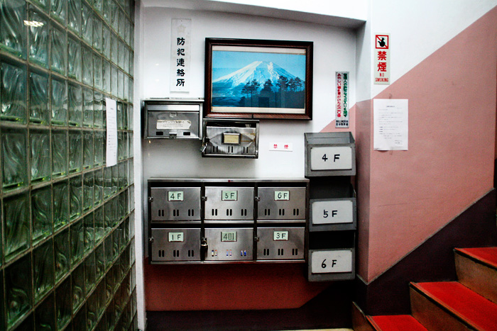 Una entrada a un edifici de pisos, amb tot de bústies a la paret i unes escales de color vermell que pugen amunt a la dreta. A sobre de les bústies una foto del Mont Fuji emmarcada.