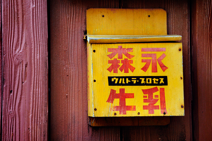 Una bústia de fusta vella, pintada de color groc sobra una paret també de fusta de color vermell terrós. La bústia té pintats uns caràcters japonesos molt grossos de color vermell sang, i la tapa està deformada per la humitat.